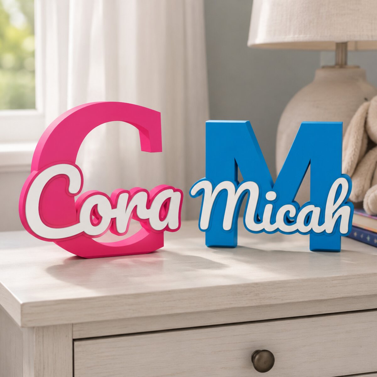 Two 3D printed name signs on a dresser: Cora in pink with a white script name, and Micah in blue with a white script name